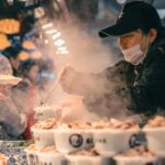 Bustling street food scene in Beijing with vendor serving steamed dishes to customers in the night market.