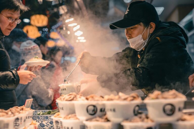 Bustling street food scene in Beijing with vendor serving steamed dishes to customers in the night market.