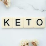Scrabble tiles spelling 'Keto' on a marble surface with white flowers, symbolizing a ketogenic lifestyle.