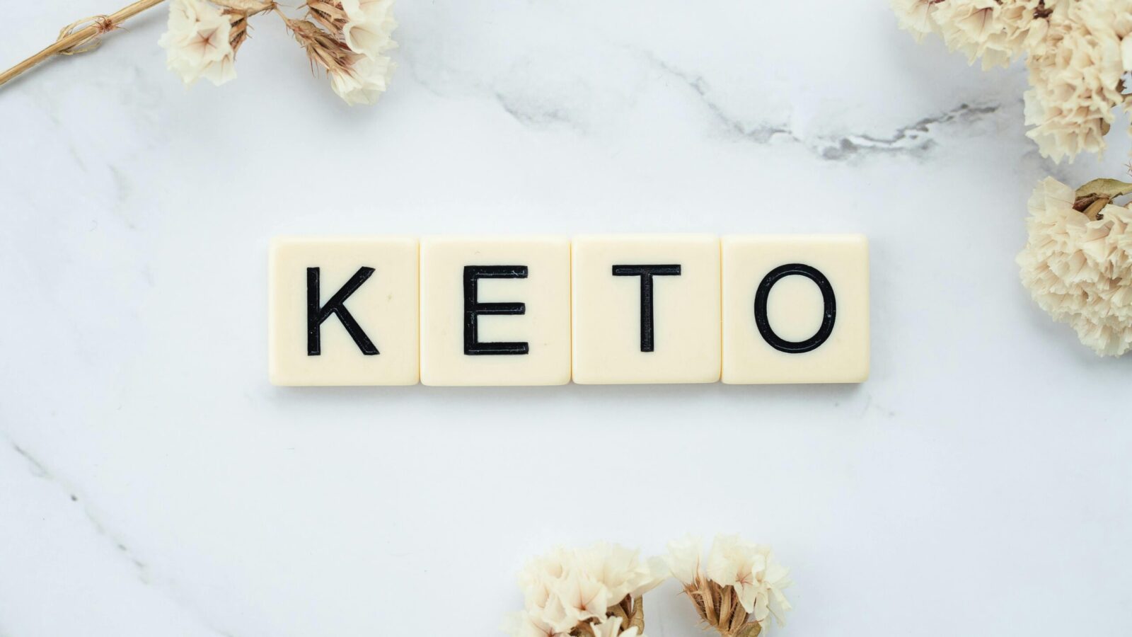 Scrabble tiles spelling 'Keto' on a marble surface with white flowers, symbolizing a ketogenic lifestyle.
